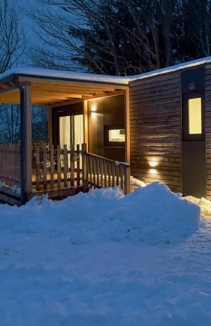 Piccola casa di legno illuminata, circondata da neve e alberi.