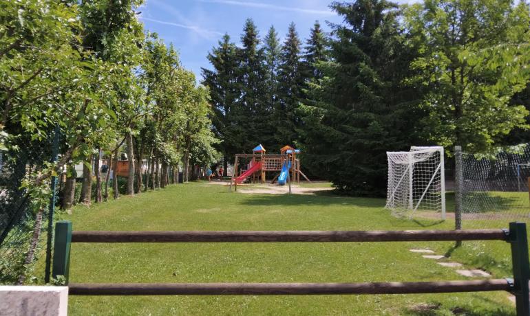 Parco giochi con scivolo, altalena e campo da calcio, alberi intorno.