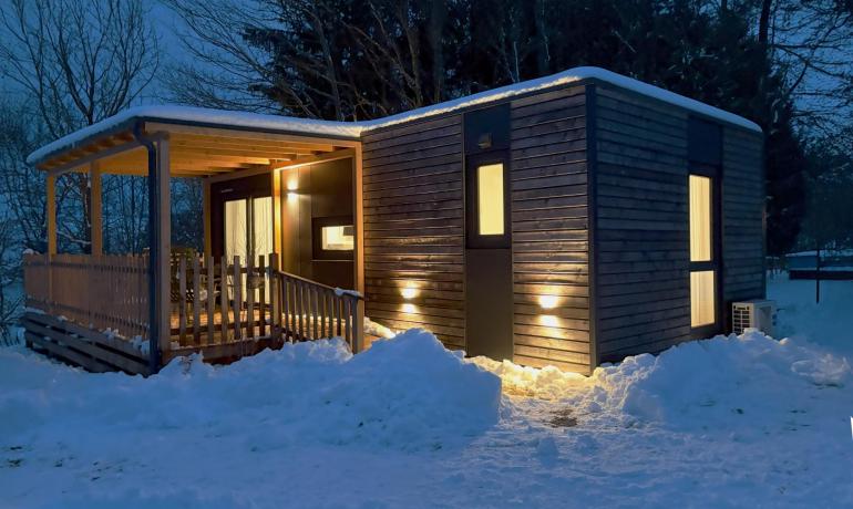 Piccola casa di legno illuminata, circondata da neve e alberi.