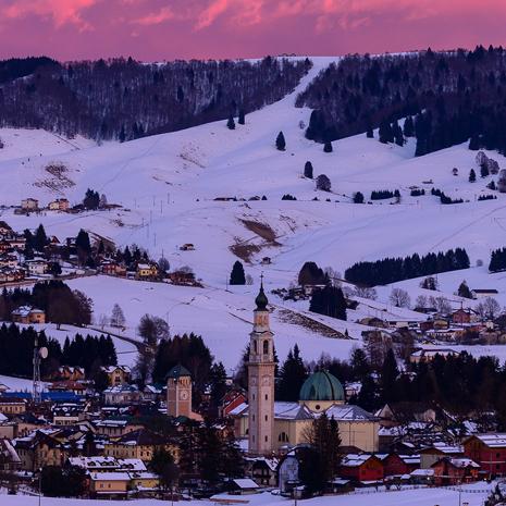 Paesaggio invernale con chiesa, montagne innevate e cielo rosa al tramonto.