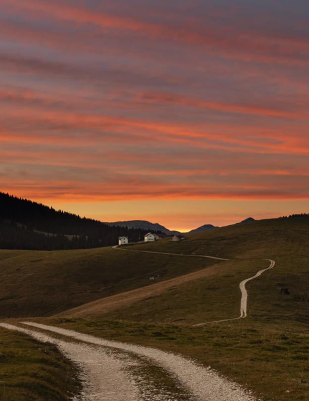 Sentiero serpeggiante tra colline al tramonto con cielo arancione.